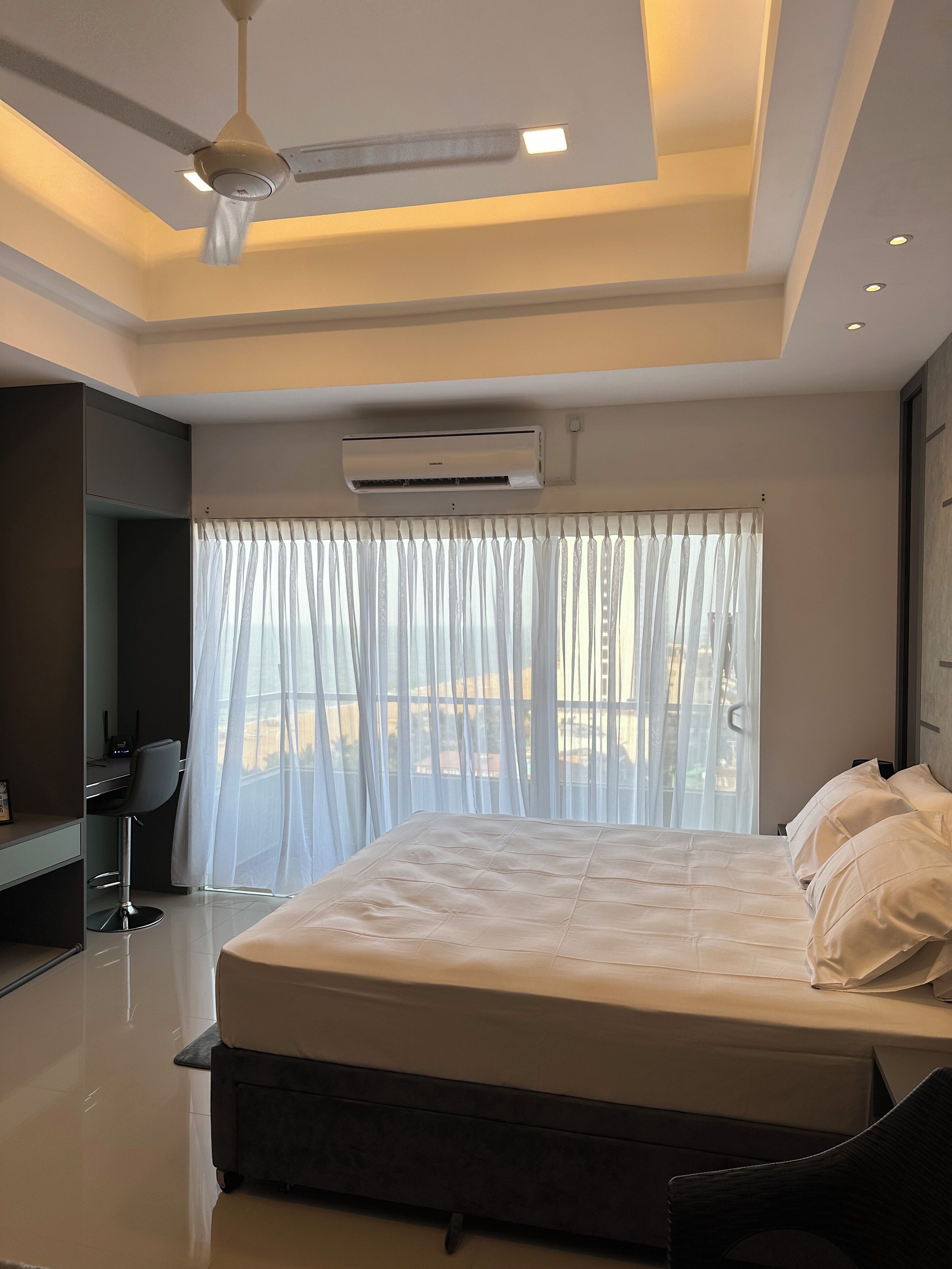 A modern bedroom with a large bed, an air conditioning unit, a ceiling fan, and a view through sheer curtains.
