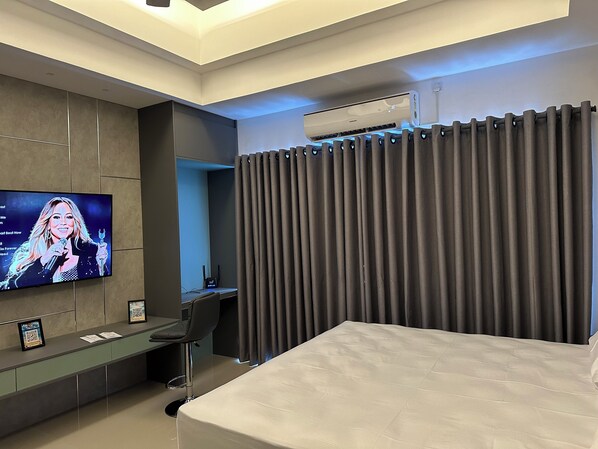 Una habitación de hotel moderna con una cama grande, un televisor de pantalla plana montado en la pared, un escritorio con silla y una ventana con cortinas oscuras.