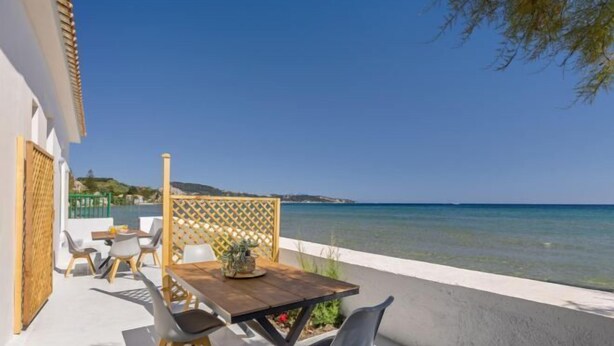 A terrace with a wooden table and chairs, a white wall, and a view of the sea. - Aliki's Summerhouse 2 (Argassi)