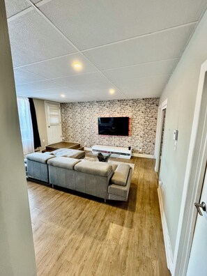 A modern living room with a stone wall, a grey sectional sofa, a flat-screen TV mounted on the wall, and a wooden floor. - Ideal stay, 3 ½-room apartment in the heart of downtown Trois-Rivières (Trois-Rivières)