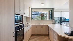 A modern kitchen with a built-in oven, microwave, and sink. It features light wood cabinetry and a large window offering a view of a cityscape and palm trees. - Family Fun at Sun City 3BR Near Beach - CoastHM (Surfers Paradise)