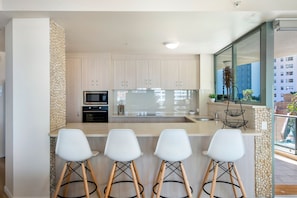 A modern kitchen with a central island, bar stools, and a large window overlooking a cityscape. - Family Fun at Sun City 3BR Near Beach (Surfers Paradise)