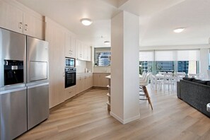 Une cuisine moderne équipée d’appareils électroménagers en acier inoxydable, un coin repas avec des meubles blancs et une vue sur la ville grâce à de grandes fenêtres.