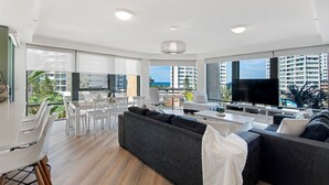 A modern living room with a large TV, a dining area, and a balcony with a view of the city and ocean. - Family Fun at Sun City 3BR Near Beach - CoastHM (Surfers Paradise)