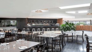 A modern restaurant with multiple dining tables and chairs, a bar area with shelves of bottles, and large windows providing natural light. - Family Fun at Sun City 3BR Near Beach - CoastHM (Surfers Paradise)