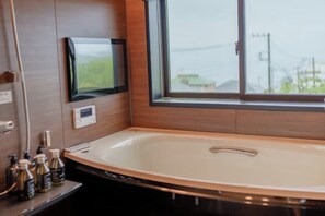 A modern bathroom with a large bathtub, a television mounted on the wall, and a window providing a view of the outdoors. - Villa Atami (Atami)