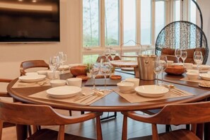 A round dining table set with plates, glasses, and cutlery, surrounded by wooden chairs in a well-lit room with a large window and a hanging chair. - Villa Atami (Atami)