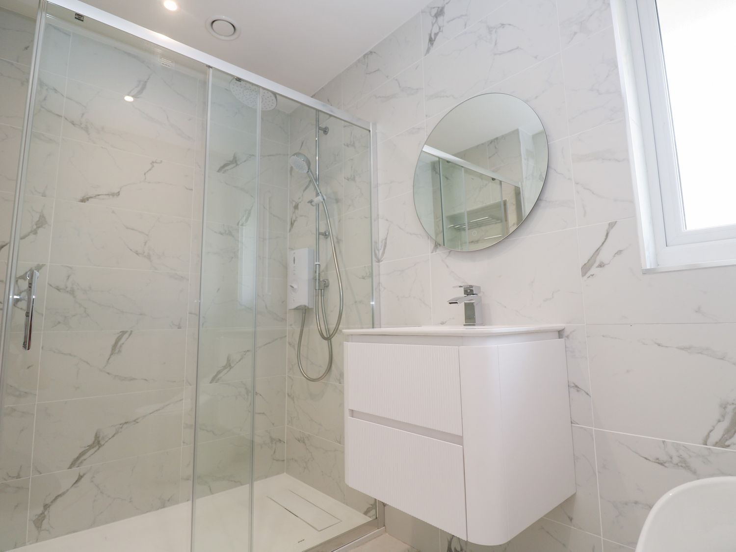 Un baño moderno con ducha envuelta en vidrio, un tocador blanco con espejo y paredes con estampado de mármol.