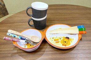 Une table ronde en bois, sur laquelle se trouvent une tasse noire, deux bols aux motifs colorés, des baguettes, une cuillère et une fourchette.
