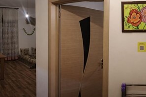 A room with a wooden door, a bed with a patterned bedspread, a wall-mounted artwork, and a QR code.