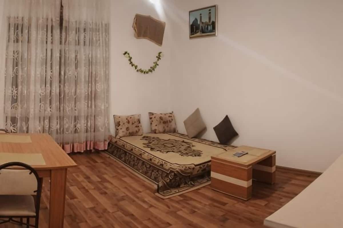 A room with a bed, a small table, a chair, and a window with sheer curtains.