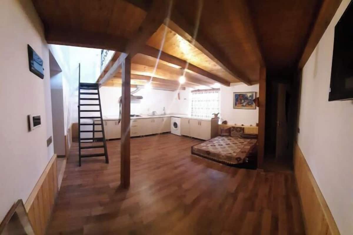A loft-style room with wooden flooring, a loft bed, a ladder, and a kitchen area.