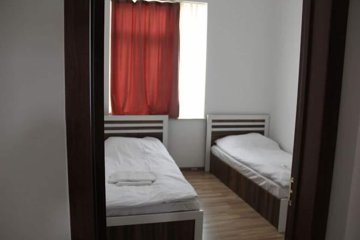 A small, narrow room with two single beds, a window with red curtains, and a wooden floor.