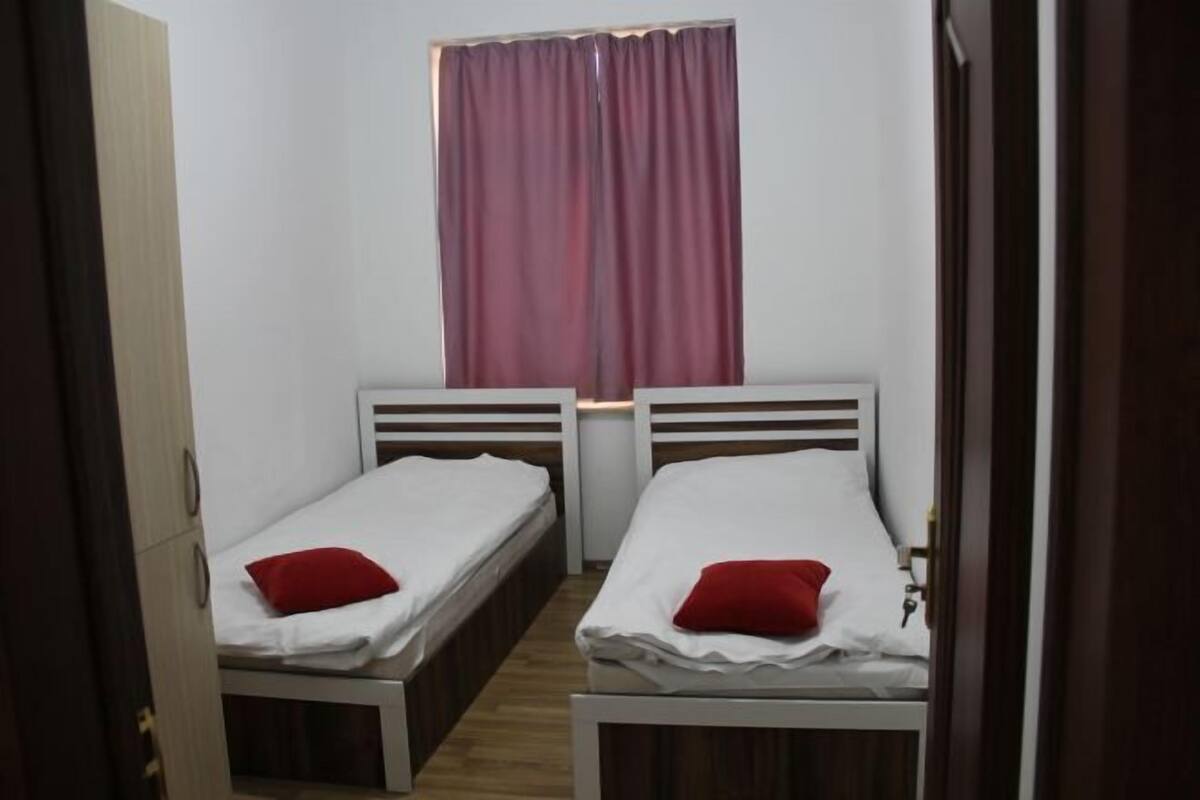 A small room with two single beds, a window with curtains, and a wooden floor.