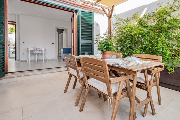 An outdoor dining area with a wooden table and chairs, a potted plant, and a view of a building with green shutters.