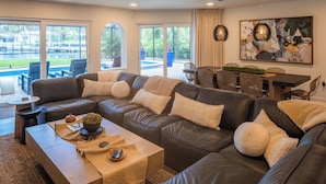 A modern living room with a sectional sofa, a dining table with chairs, and a large window with a view of a pool and greenery.