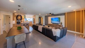 A modern living room with a large TV, a sofa, a dining area, and a kitchen in the background.