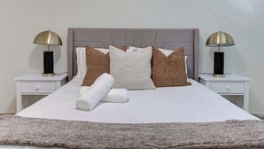 Cama bem arrumada com cabeceira cinza, roupa de cama branca e toalha branca enrolada. Há duas luminárias de cabeceira com base preta e cúpulas douradas.