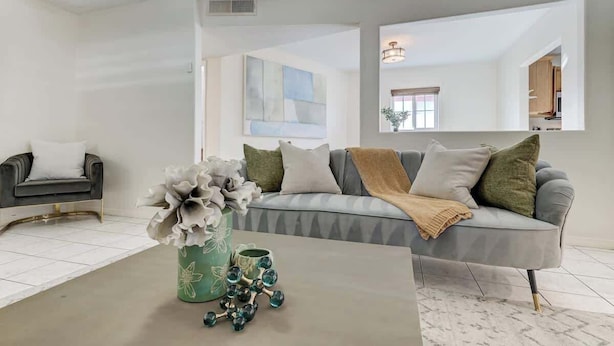 Uma sala de estar moderna com um sofá cinzento, uma mesa de centro com objetos decorativos e um espelho grande que reflete o ambiente.