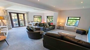 A spacious living room with a large sofa, a smaller sofa, and a coffee table. There are large windows with curtains, a radiator, and a lamp on a side table. - Streamways Slps 12-16 Hot Tub with Views & Cabin (Croyde)