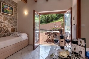 Een kamer met een bed, een eettafel met wijnglazen en flessen, en een open deur naar een buitengebied met een tafel en stoelen.