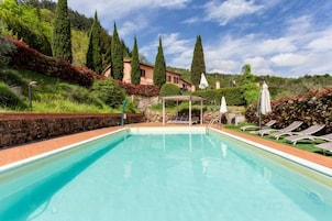 Una piscina con sillones de descanso y una pérgola, rodeada de jardines ajardinados y una colina.