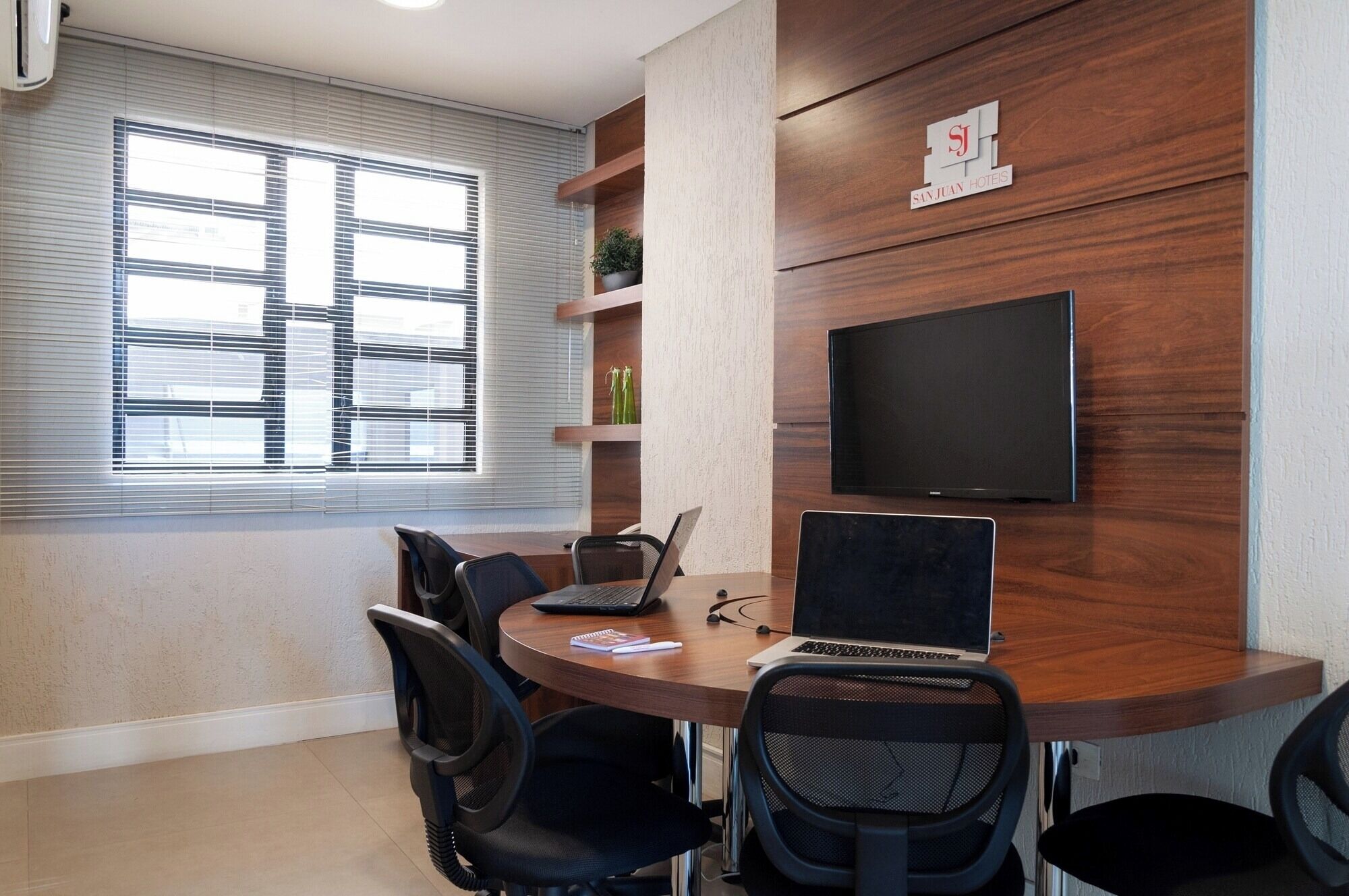 A conference room with a round table, four chairs, a TV mounted on the wall, and a window with blinds.