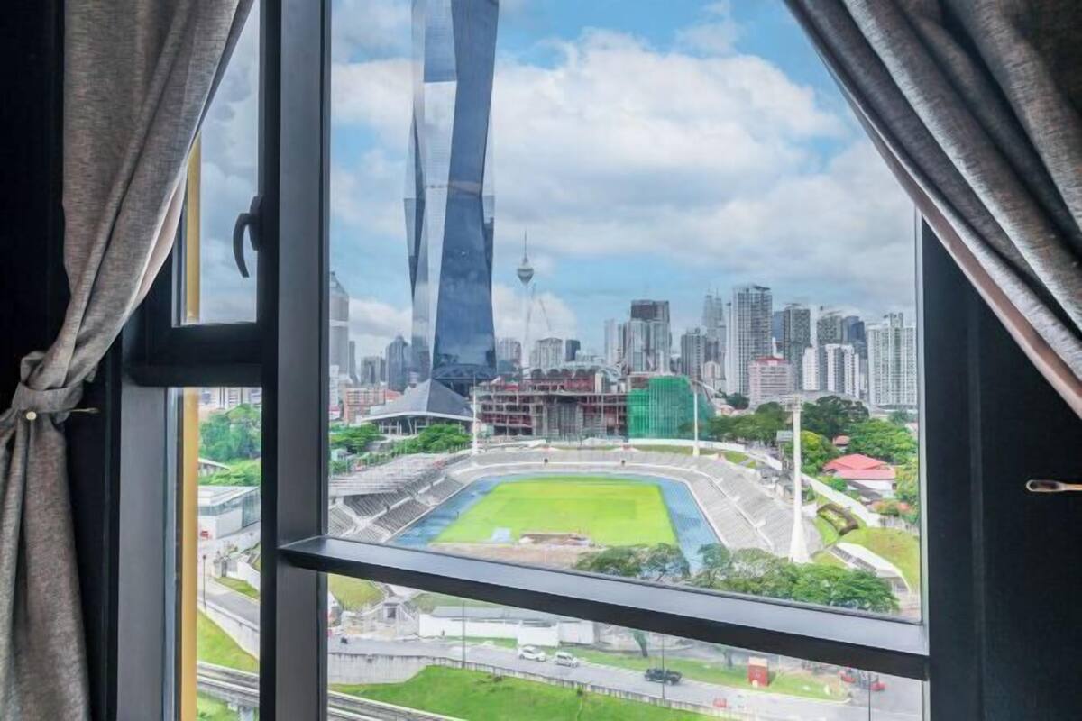 Blick aus einem Fenster mit Vorhängen, der eine Stadtansicht mit einem hohen Wolkenkratzer, einem Stadion und umliegenden Gebäuden zeigt.