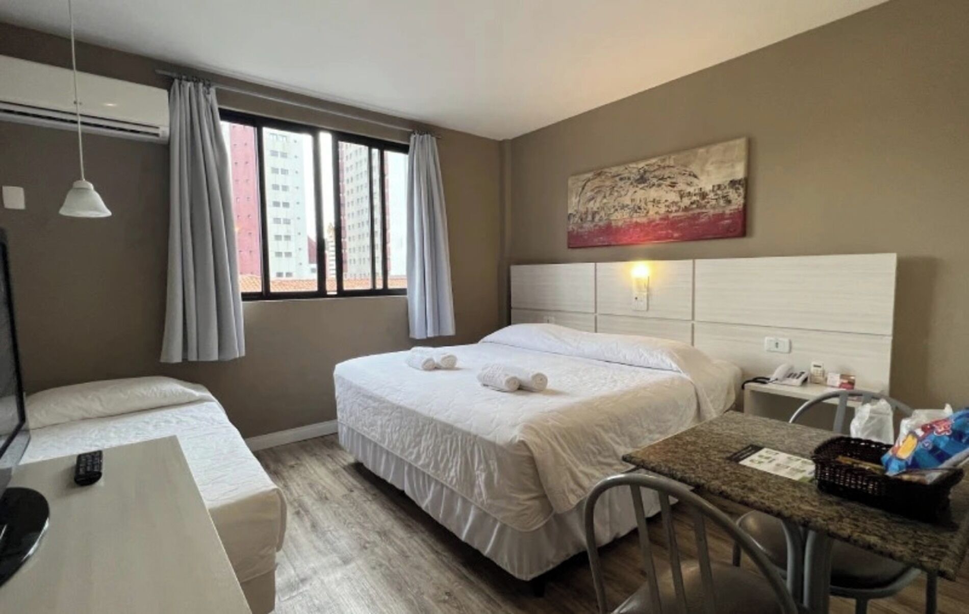 A hotel room with a bed, a desk, a chair, a table, a telephone, and a painting on the wall.