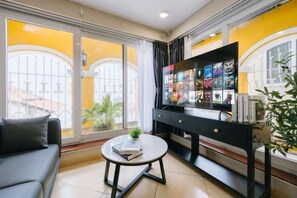 A living room with a sofa, coffee table, TV stand with a flat-screen TV, and a large window with yellow curtains. - VietHOME - 22 Pho Hue - Duplex 67 (Hanoi)