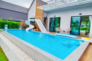 A modern outdoor swimming pool with a slide, a lounge area with chairs, and a glass door leading to a covered patio. - Relax Pool Villa Khaoyai (Pak Chong)