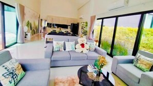 A modern living room with a grey sofa, a large teddy bear, and a dining area in the background. - Relax Pool Villa Khaoyai (Pak Chong)