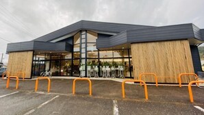 Um edifício moderno com uma ampla entrada de vidro, painéis de madeira nas laterais e guarda-corpos laranja na área de estacionamento.