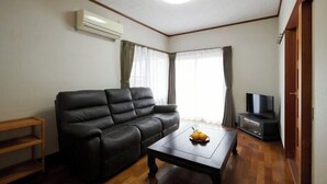 Uma sala de estar com um sofá de couro preto, uma mesa de centro de madeira com frutas, um rack para TV e um ar condicionado.