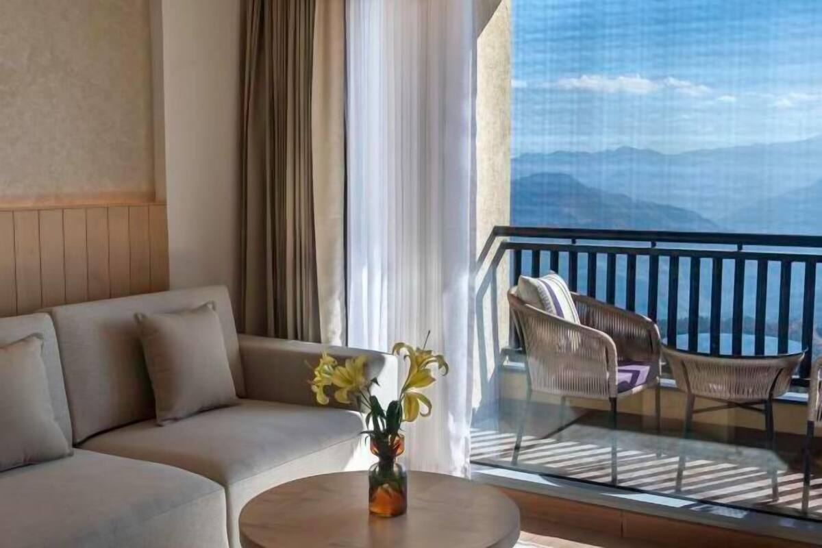A modern living room with a sofa, a round coffee table with a vase of flowers, and a balcony with wicker chairs and a view of mountains.
