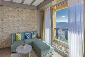 A modern living room with a stone wall, a green sofa with patterned cushions, a small round table with fruit, and a balcony with a view of mountains. - dusitD2 Fagu (Shimla)