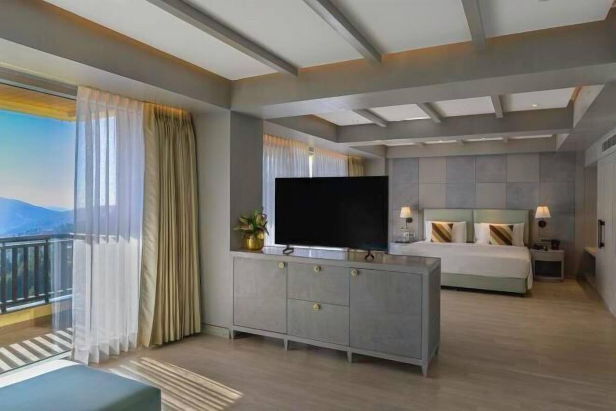 A modern hotel room with a large bed, a TV on a grey cabinet, a balcony with curtains, and a cityscape view.
