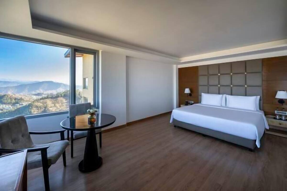 A hotel room with a large bed, a small table with a chair, a view of mountains through a large window, and a wooden floor.