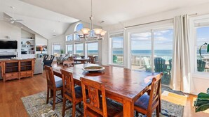 A spacious dining area with a large wooden table and chairs, a chandelier, and a view of the ocean through large windows. - SS2-Oceanfront w/7 masters, private pool & hot tub, elevator & pool table (Southern Shores)