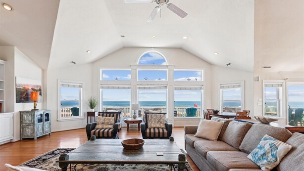A spacious living room with a high ceiling, large windows offering ocean views, a ceiling fan, and a cozy seating area with a coffee table. - SS2-Oceanfront w/7 masters, private pool & hot tub, elevator & pool table (Southern Shores)