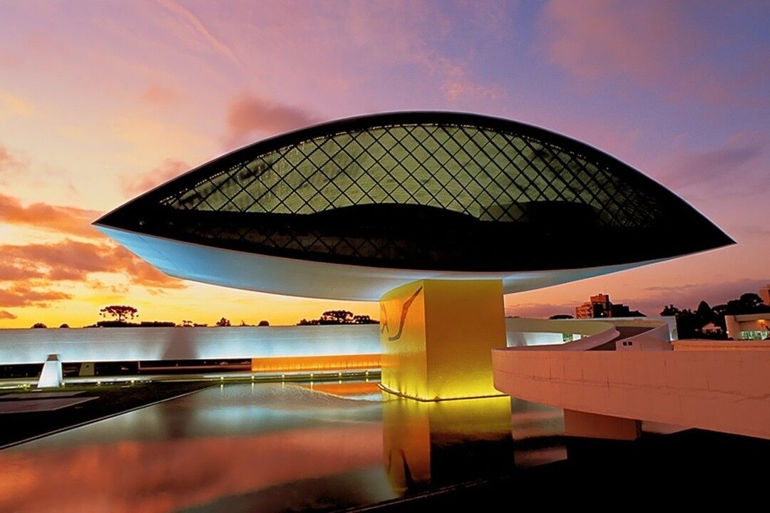 A modern architectural structure with a unique, curved roof design, situated in an open area with a reflective water surface in the foreground.