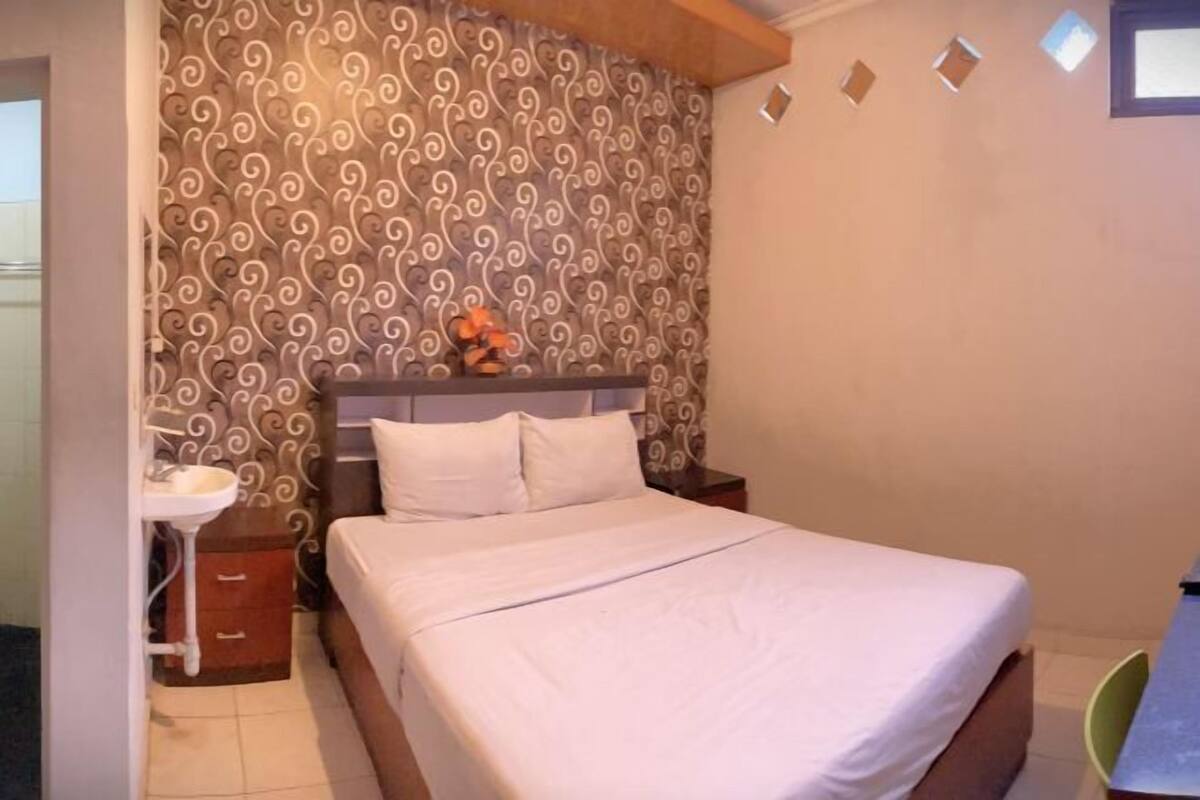 Ein Schlafzimmer mit einem Bett, einem Nachttisch, einem Wandregal mit Dekoration und einem Fenster mit Jalousien.