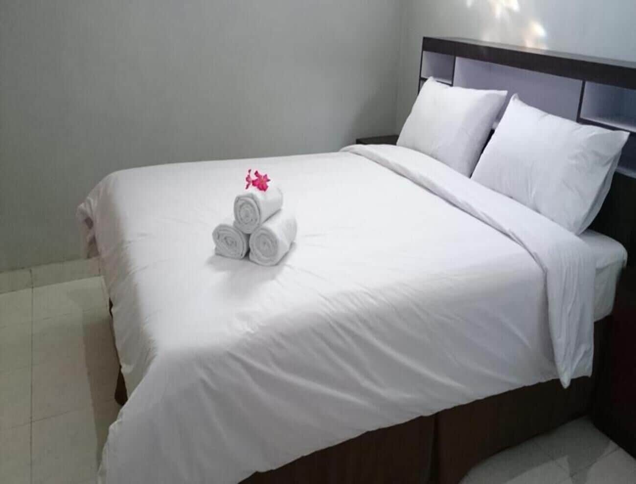 A neatly made bed with white linens and a rolled towel with a pink flower.