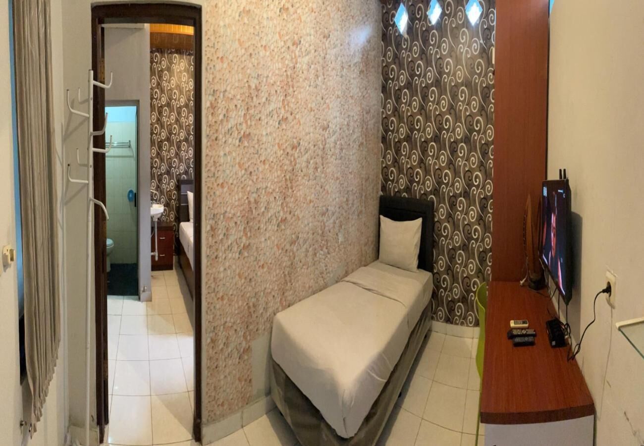 Une petite chambre avec un lit, une table de chevet sur laquelle se trouvent une télévision et des télécommandes, une salle de bain visible à travers une porte ouverte, et un mur orné d’un motif.