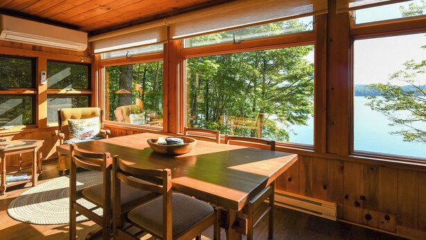 Una cabaña de madera con comedor, ventanas amplias y vistas a un lago.