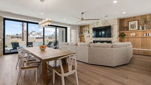A modern living room with a wooden dining table, beige sofa, and a flat-screen TV mounted on a stone wall. - Lula Bay Lagoon House: Private Pool + Near Lagoon (St. George)