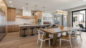 A modern kitchen with wooden cabinets, a central island with bar stools, and a dining area with a wooden table and white chairs. - Lula Bay Lagoon House: Private Pool + Near Lagoon (St. George)