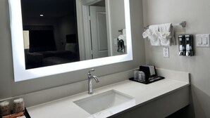 A hotel bathroom with a large mirror, a sink, a towel rack, and a soap dispenser. - Hotel Royale (Houston)