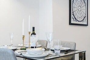 Una mesa de comedor dispuesta con platos blancos, vasos transparentes y una botella de vino, con una obra de arte abstracta enmarcada en la pared.