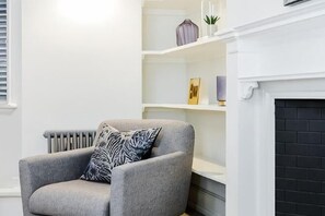 Un salón con un sillón gris, un cojín blanco y negro estampado, una chimenea blanca y estanterías con objetos decorativos.